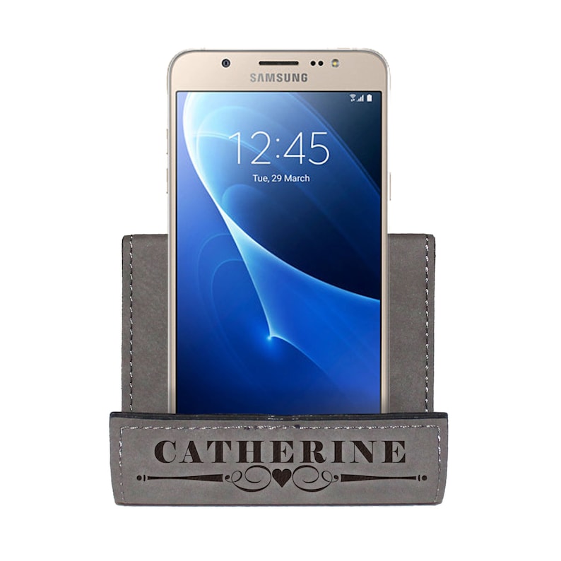 May include: A gray leather phone stand with a personalized inscription that reads "CATHERINE" with a heart design. A gold Samsung phone is in the stand.