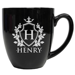 Pu&ograve; includere: Una tazza in ceramica nera lucida con un disegno stampato in bianco sulla parte anteriore. Il design include uno scudo con la lettera H all'interno, sormontato da una corona e fiancheggiato da rami frondosi. Il nome HENRY &egrave; stampato in bianco.