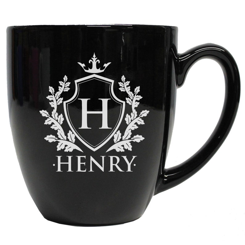Pu&ograve; includere: Una tazza in ceramica nera lucida con un disegno stampato in bianco sulla parte anteriore. Il design include uno scudo con la lettera H all'interno, sormontato da una corona e fiancheggiato da rami frondosi. Il nome HENRY &egrave; stampato in bianco.