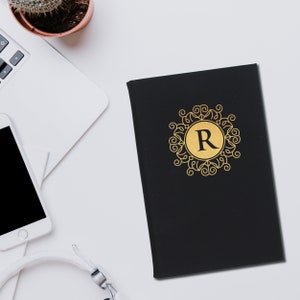 May include: Black journal with a gold monogram letter "R" inside a decorative emblem. The journal is on a white surface, next to a laptop, a smartphone, and headphones. A small potted cactus is also visible.