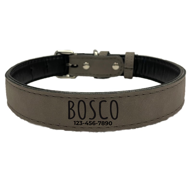 Op de afbeelding: Een grijze hondenriem met de naam "BOSCO" en een telefoonnummer in zwarte letters. De halsband heeft een zilveren gesp en klinknagels. De binnenkant van de halsband is zwart.