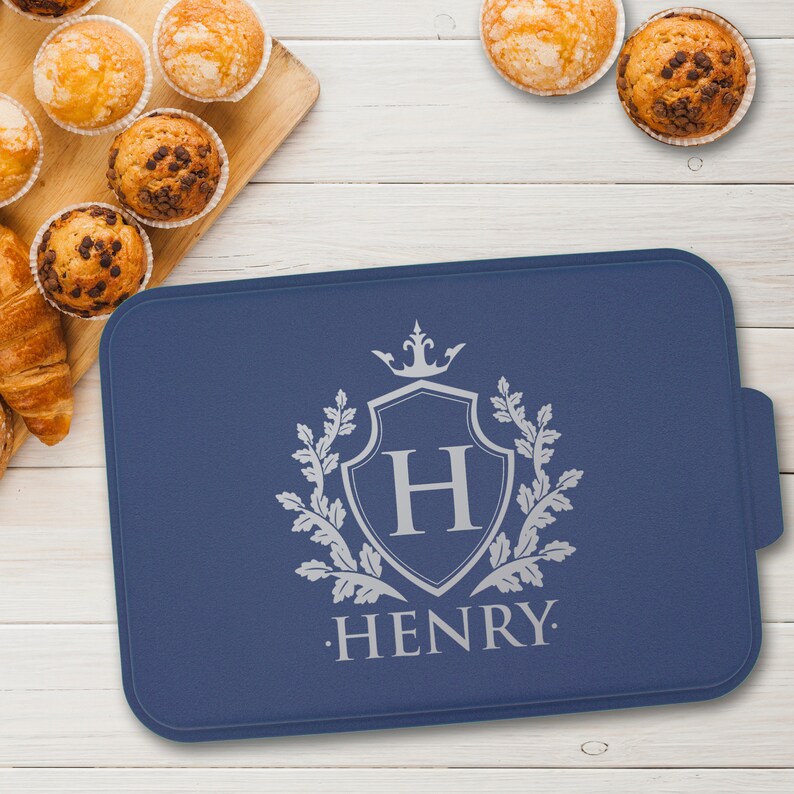 May include: A blue silicone baking mat with a silver design of a shield with the letter H and a crown above it. The text "HENRY." is below the shield.