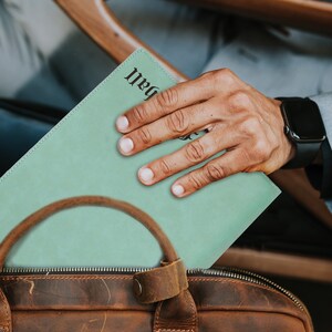 May include: A light green leather portfolio with the text "ball" in a black gothic font, partially inserted into a brown leather briefcase with a rounded handle and a metal snap closure, featuring visible stitching along the edges.