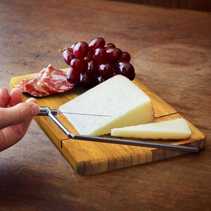 May include: A bamboo cheese board with a built-in cheese knife. The board has a groove for cutting cheese and a section for serving. There is a block of white cheese, a wedge of cheese, and a few slices of salami on the board. There are also red grapes on the board.