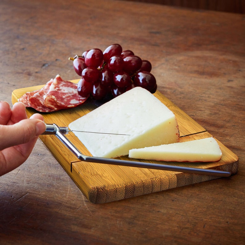 May include: A bamboo cheese board with a built-in cheese knife. The board has a groove for cutting cheese and a section for serving. There is a block of white cheese, a wedge of cheese, and a few slices of salami on the board. There are also red grapes on the board.