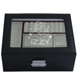 May include: A black rectangular jewellery box with a clear glass lid featuring a white engraved graphic of a unicorn surrounded by stars and the name IZZY. The interior is lined with light grey fabric, including ring rolls and compartments.