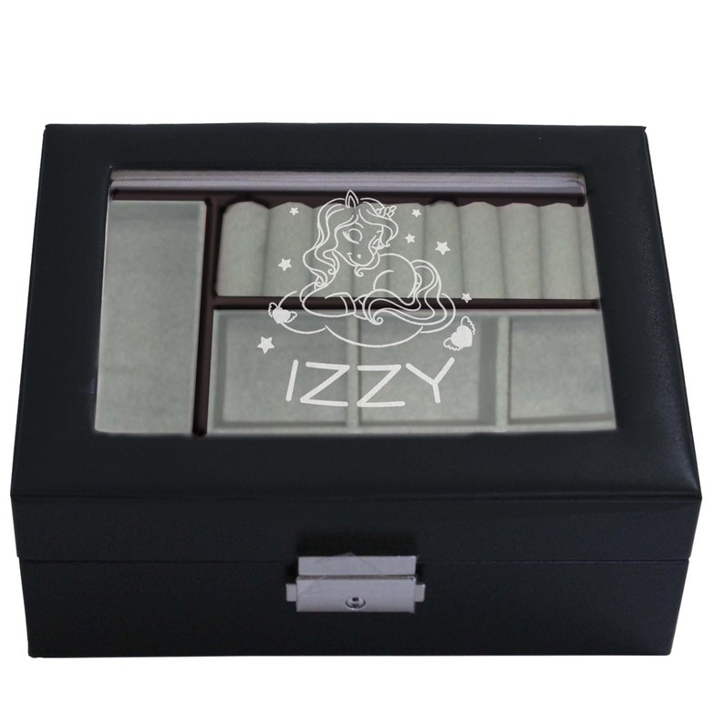 May include: A black rectangular jewellery box with a clear glass lid featuring a white engraved graphic of a unicorn surrounded by stars and the name IZZY. The interior is lined with light grey fabric, including ring rolls and compartments.