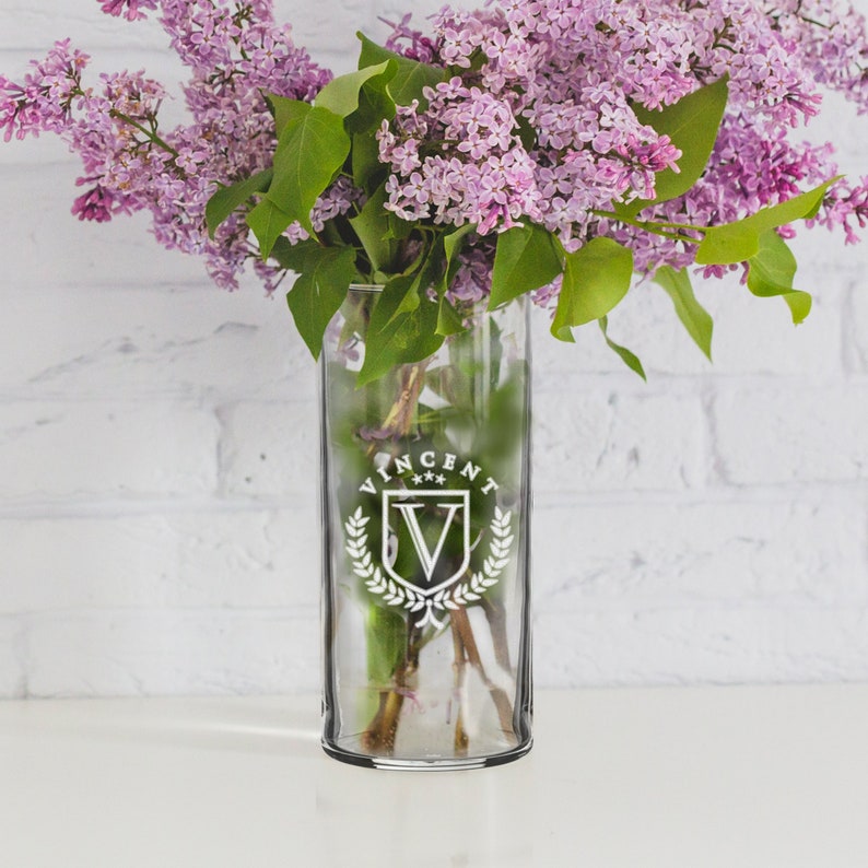 May include: A clear glass vase with a white emblem that reads "VINCENT" and a "V" inside a crest. The vase holds a bouquet of purple lilacs with green leaves. The background is a white brick wall.