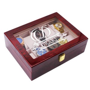 May include: A cherry-stained wooden watch box with a clear glass top. The box is divided into compartments, each holding a watch. The name "DRAKE" is displayed in white script on the inside of the lid. A gold-colored clasp secures the box.