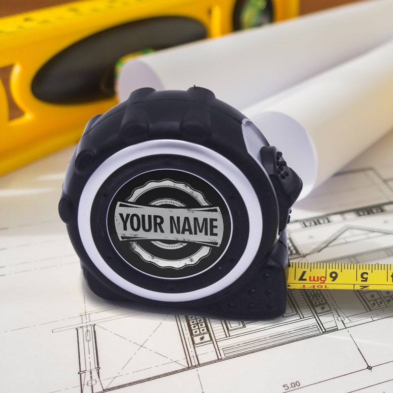 May include: A black tape measure with the text "YOUR NAME" on the face, resting on a blueprint. A yellow tape extends from the measure, showing measurements in inches. A yellow level and rolled blueprints are in the background.
