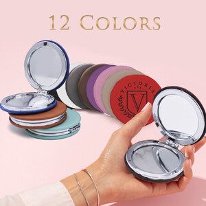 May include: A collection of round compact mirrors in 12 colors, featuring silver-toned metal frames and hinges. One red mirror displays the text VICTORIA with a shield and laurel wreath logo. The mirrors are shown both stacked and held open.