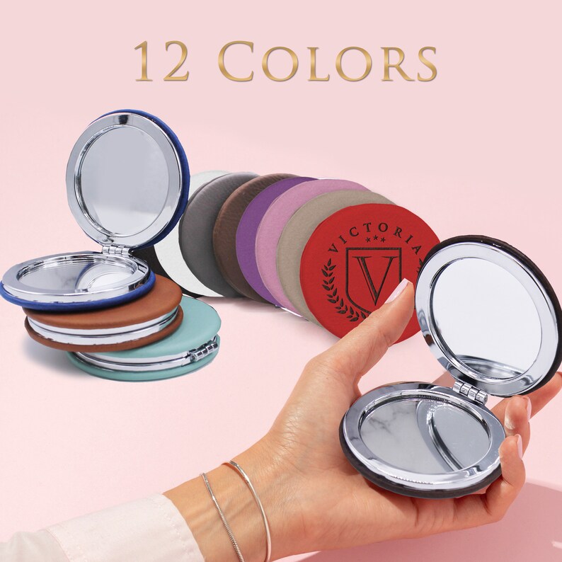 May include: A collection of round compact mirrors in 12 colors, featuring silver-toned metal frames and hinges. One red mirror displays the text VICTORIA with a shield and laurel wreath logo. The mirrors are shown both stacked and held open.