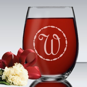 May include: A clear stemless wine glass filled with deep red liquid, featuring an etched circular border containing the letter W in a script font. A white carnation flower with a dark red ribbon is positioned at the base of the glass.