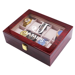 Pode incluir: Uma caixa de rel&oacute;gios de madeira castanho-avermelhada escura com tampa de vidro, contendo v&aacute;rios rel&oacute;gios. A caixa tem um fecho dourado e o nome "JAMES" est&aacute; impresso no vidro. A caixa &eacute; dividida em compartimentos.
