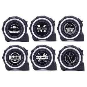 May include: Six black and white personalized tape measures. Each tape measure has a white circular face with a different name or text, such as "Worlds Best Dad Henry" or "Miller". The tape measures are for construction or DIY projects.