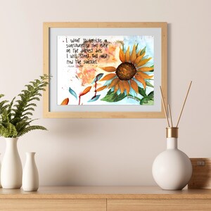 May include: Watercolor painting of a sunflower in a wooden frame. The artwork features an orange sunflower with green leaves and the quote "I want to be like a sunflower..." in black text. The background is light blue.
