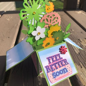 May include: A handmade pop-up card with a floral arrangement. The card features green, orange, and white paper flowers with yellow gem accents. The front of the card reads "Feel Better Soon" with a bandage illustration.