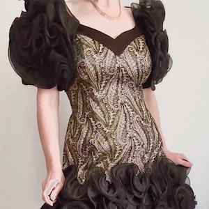 May include: A vintage cocktail dress with a gold and black paisley pattern bodice and a black ruffled skirt. The dress features dramatic black ruffled sleeves and a V-neckline. The dress is a classic example of vintage evening wear.