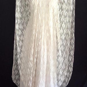 May include: Ivory lace dress with a V-neck and flowing sleeves. The dress has a layered design, with a sheer lace overlay over a solid fabric. The dress is floor-length and suitable for special occasions.