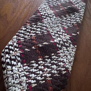 Op de afbeelding: Een geweven stropdas met een textuurpatroon. De stropdas heeft een mix van bruine, witte en roze draden, waardoor een gedetailleerd ontwerp ontstaat. De stropdas wordt getoond op een houten oppervlak.