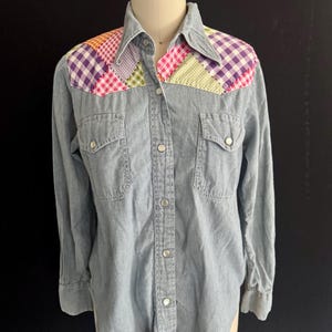 Könnte beinhalten: Hellblaues Jeanshemd mit bunten Patchwork-Akzenten auf den Schultern. Das Patchwork umfasst rosa, lila, grüne und orangefarbene Karos und gemusterte Stoffe. Mit langen Ärmeln, zwei Brusttaschen und silbernen Druckknöpfen.