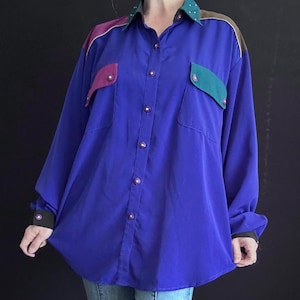 May include: A long-sleeved, button-down shirt in a vibrant blue. The shirt features color-blocked accents in purple, teal, and brown on the shoulders and pockets. The collar has white polka dots. The cuffs are black.