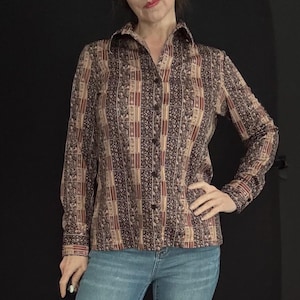 May include: A button-down shirt with a brown, tan, and burgundy geometric pattern. The long-sleeved shirt has a collared neckline and is paired with blue jeans. The shirt's pattern is a repeating design of rectangles and floral elements.