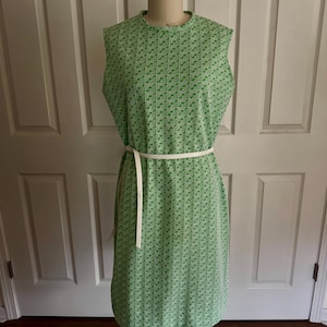 May include: A sleeveless, knee-length green dress with a geometric pattern. The dress has a high neckline and is cinched at the waist with a white belt. The dress is displayed on a mannequin.