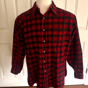 Pode incluir: Uma camisa de flanela xadrez vermelha e preta com mangas compridas e fecho de botões. A camisa é exibida em um manequim. O padrão xadrez inclui detalhes verdes. A camisa tem gola e bolso no peito.