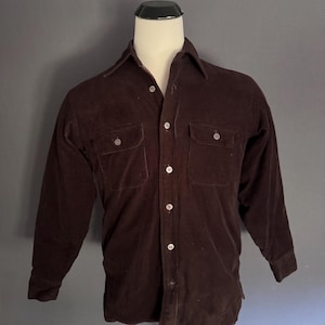 May include: A long-sleeve dark brown corduroy shirt featuring a standard collar, a full-length button-down front closure, and two chest pockets with buttoned flaps. The garment is displayed on a mannequin, showcasing its textured fabric.
