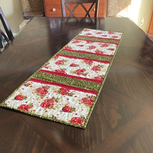 May include: A green and red table runner with a pomegranate pattern. The runner is quilted and has a white background.