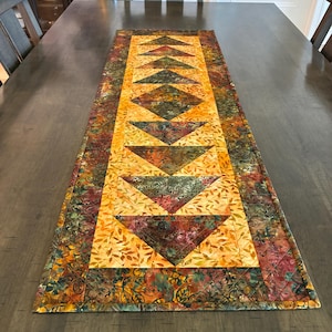 May include: A rectangular quilted table runner featuring a central row of dark green and burgundy triangles set against a golden yellow background with a leaf pattern. The border consists of a multi-colored fabric with a dark floral motif.