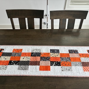May include: A rectangular quilted table runner featuring a patchwork design of alternating squares in orange, black, white, and gray. The fabric squares display various Halloween-themed patterns including pumpkins, skeletons, and spiderwebs.