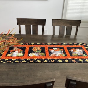 May include: A black quilted table runner with four orange and black squares featuring a fall harvest theme. Each square has a basket of pumpkins, gourds, and sunflowers. The runner is on a dark wood table.