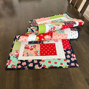 May include: Three colorful quilted table runners with floral and geometric patterns. The runners feature red, pink, green, and navy blue fabrics. The table runners are folded and stacked on a dark wooden table.