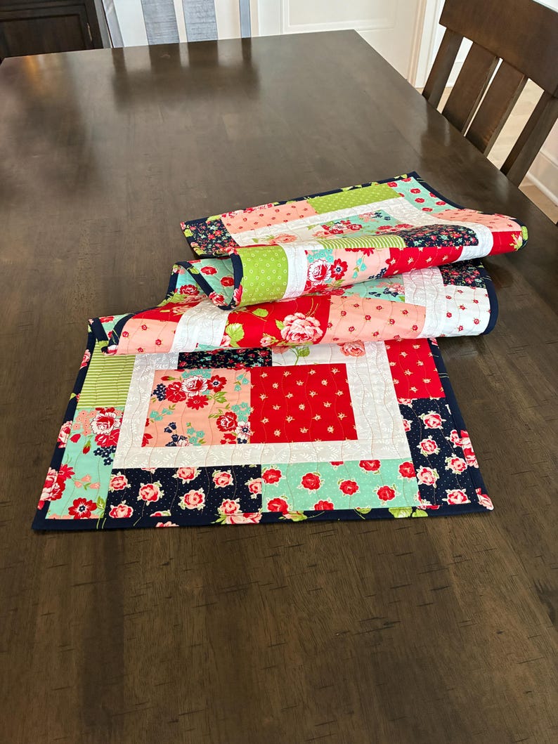 May include: Three colorful quilted table runners with floral and geometric patterns. The runners feature red, pink, green, and navy blue fabrics. The table runners are folded and stacked on a dark wooden table.
