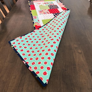 May include: A handmade table runner with a patchwork design on one side and a floral pattern on the other. The patchwork features squares of red, green, and floral fabrics. The reverse side has a turquoise background with red flower prints.