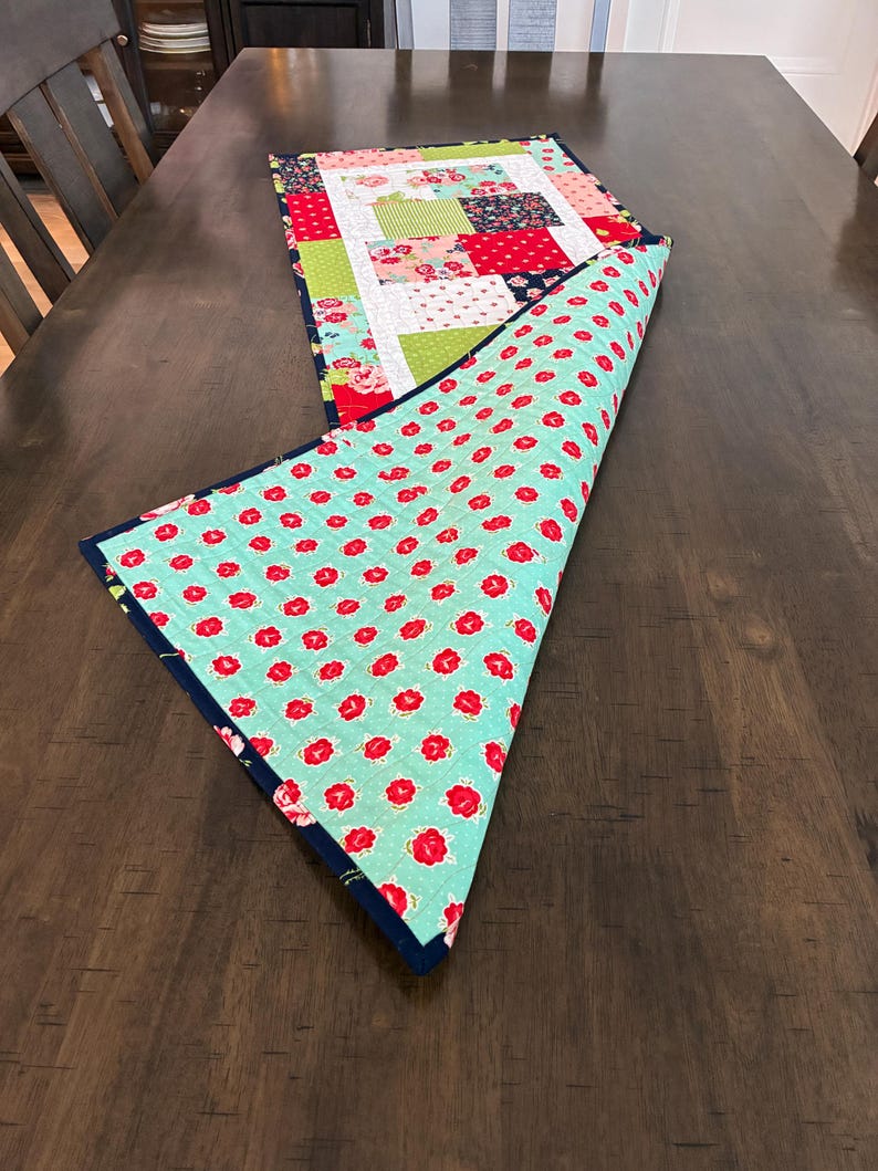 May include: A handmade table runner with a patchwork design on one side and a floral pattern on the other. The patchwork features squares of red, green, and floral fabrics. The reverse side has a turquoise background with red flower prints.