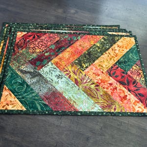 May include: A set of rectangular fabric placemats featuring a diagonal chevron pattern. The design consists of quilted strips in autumn-inspired colors including deep red, burnt orange, golden yellow, and forest green, finished with a dark green border.
