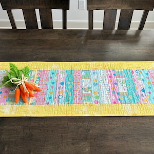 May include: A colorful Easter-themed table runner with a yellow border. The center features vertical stripes of pastel colors and Easter-related patterns. A bundle of orange carrots with green leaves is tied with twine.