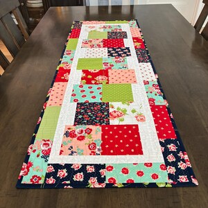 May include: A rectangular table runner with a patchwork design. The runner features squares of floral and patterned fabrics in red, pink, green, and navy blue, bordered by a navy blue fabric. The runner is placed on a dark brown wooden table.
