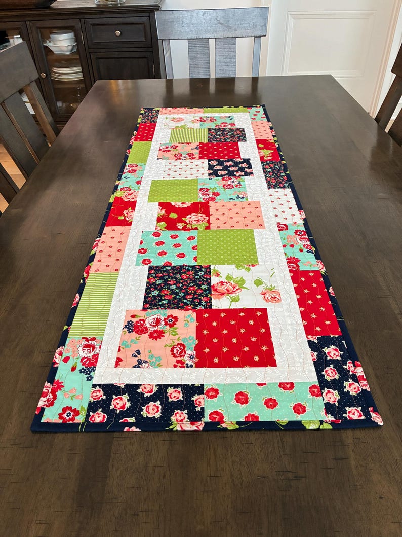 May include: A rectangular table runner with a patchwork design. The runner features squares of floral and patterned fabrics in red, pink, green, and navy blue, bordered by a navy blue fabric. The runner is placed on a dark brown wooden table.