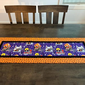 May include: A rectangular fabric table runner featuring a central panel with a purple background, haunted house illustrations, and yellow moons. The center is framed by a black border and an outer orange border patterned with small black bats.
