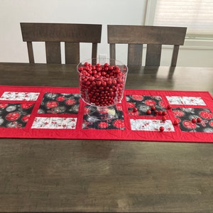 May include: A red quilted table runner with a patchwork design featuring black and white ornaments. A clear glass bowl filled with red ornaments sits on the runner.