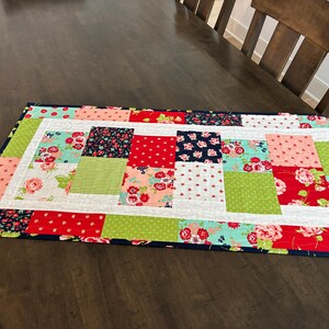 May include: A rectangular patchwork table runner with a navy blue border. The runner features squares of red, green, pink, and floral patterns. The table runner is placed on a dark brown wooden table.