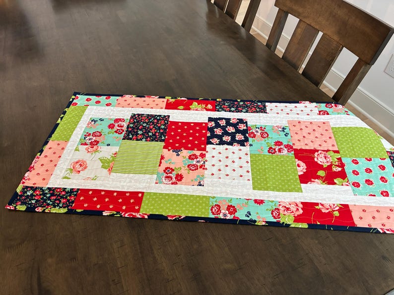 May include: A rectangular patchwork table runner with a navy blue border. The runner features squares of red, green, pink, and floral patterns. The table runner is placed on a dark brown wooden table.