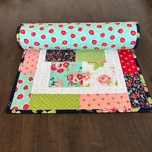 May include: A rolled-up, colorful quilt with a floral pattern. The quilt features a variety of fabrics, including a light blue fabric with red flowers, and other fabrics with floral and geometric patterns. The quilt is bordered with a dark blue fabric.