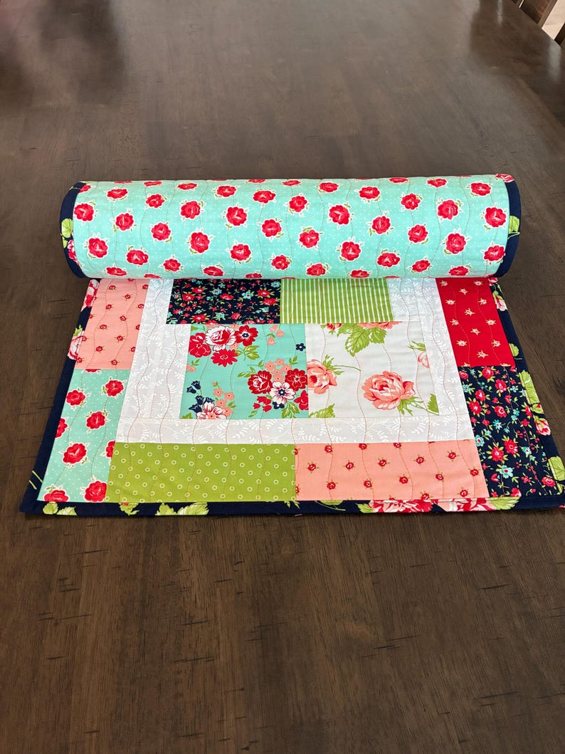 May include: A rolled-up, colorful quilt with a floral pattern. The quilt features a variety of fabrics, including a light blue fabric with red flowers, and other fabrics with floral and geometric patterns. The quilt is bordered with a dark blue fabric.