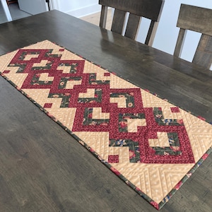 May include: A rectangular table runner with a geometric pattern. The runner features a repeating design of burgundy and floral print blocks set against a beige background. The runner is placed on a dark wooden table.