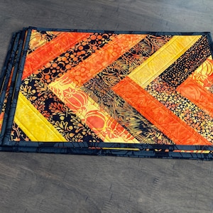 May include: Set of four quilted placemats in a chevron pattern with black, orange, and yellow fabric. The placemats have a black border.